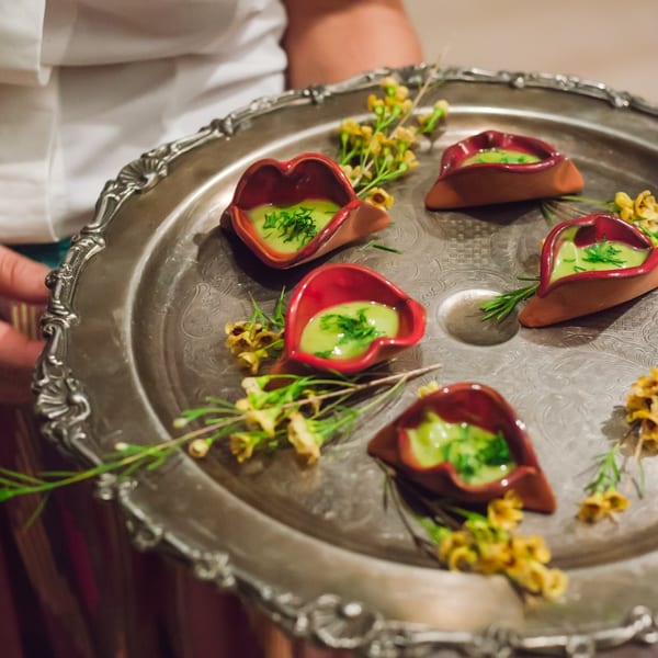 A server holds a silver tray with five small, red, lip-shaped dishes of green sauce.