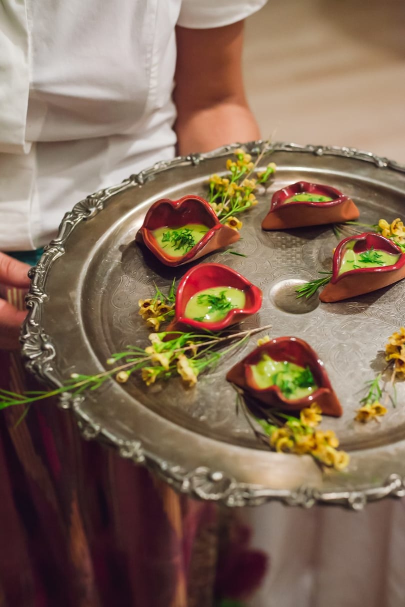 A server holds a silver tray with five small, red, lip-shaped dishes of green sauce.