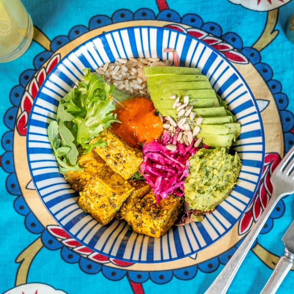 Colorful vegan bowl with tofu, avocado, rice, red cabbage, and green hummus on a patterned tablecloth.
