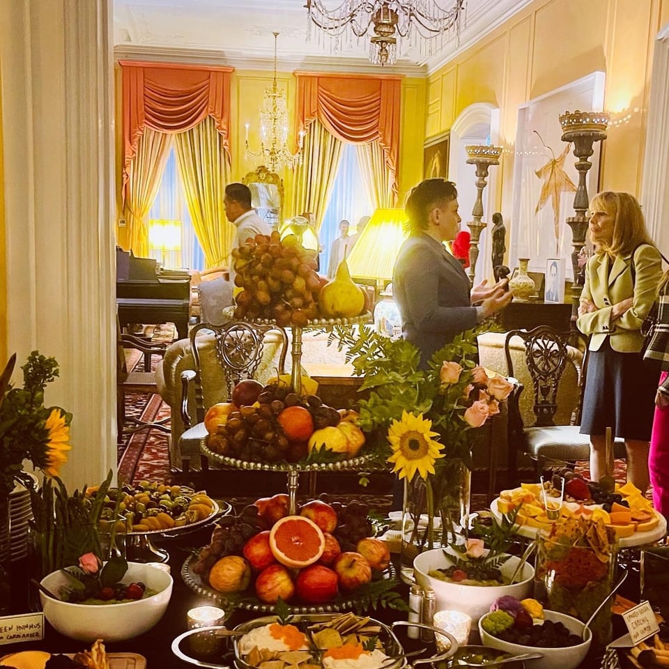 A lavish buffet table laden with fruits, cheeses, and dips, with guests mingling in a grand room.