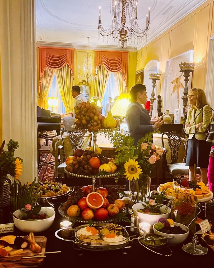 A lavish buffet table laden with fruits, cheeses, and dips, with guests mingling in a grand room.