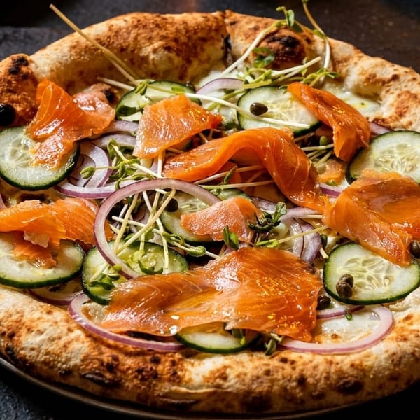 Smoked salmon pizza with cucumber, red onion, capers, and microgreens on a dark surface.