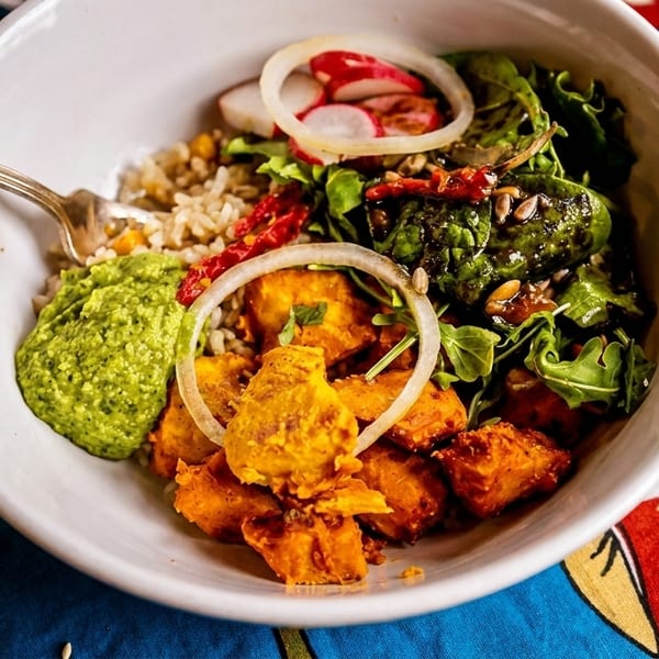 A colorful bowl of food with rice, greens, roasted sweet potatoes, and green sauce on a floral tablecloth.